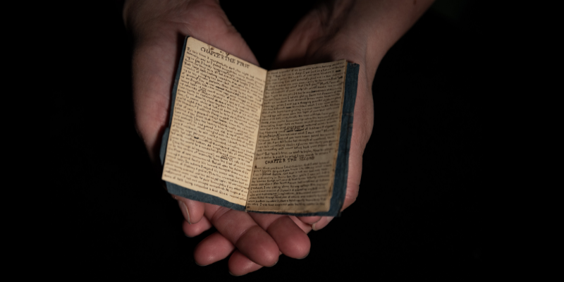 Open hands holding a tiny book filled with handwriting, against a black background.