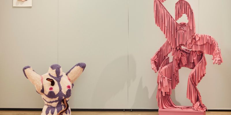 Two fabric sculptures against a white wall. On the left is a furry female torso and on the right is a figure made from wire and pink fabric fringing.