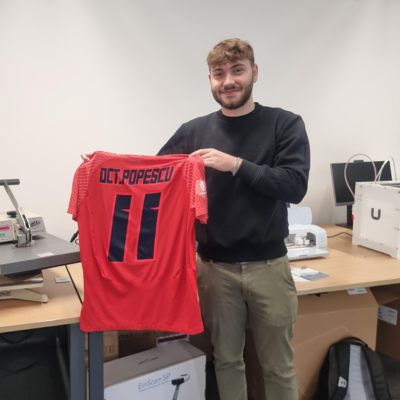 A student is standing in the Library Makerspace smiling as they hold up a red football shirt.