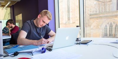 A student is sat at a desk in the Laidlaw Library. They are writing notes in front of a laptop.