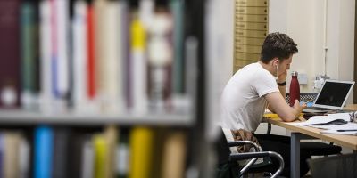 A person completing work on a laptop in the background with a bookcase in the foreground to the left