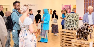 Groups of people chatting and taking pictures whilst stood among artworks in a white walled gallery. In the centre of the room are ceramics on plinths. Colourful designs hang on the far walls.