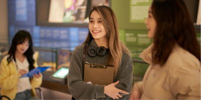 Three people in the Treasures of the Brotherton. Two are standing and smiling, one holding a clipboard. In the background, the third person is sat looking at the objects in the cases and writing on a clipboard.
