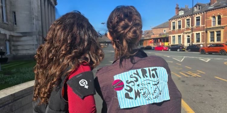 Two people stood on a street facing away from the camera with fabric protest badges affixed to their backs and sleeves.